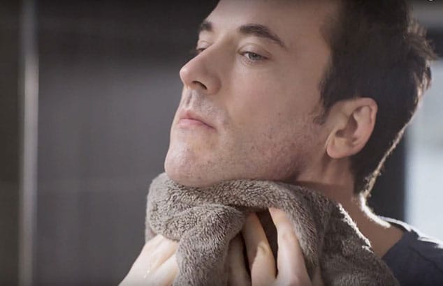 A man with light stubble and brown hair holds a brown towel against his neck.