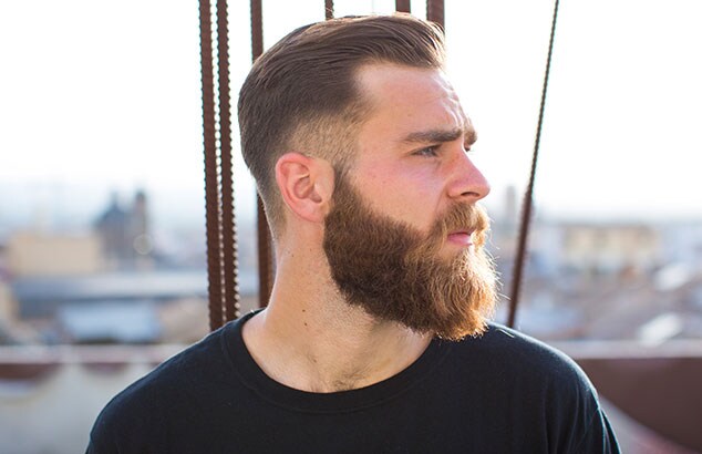 : Side profile of man with a long, brown beard and short, brown hair with a city out-of-focus in the background.
