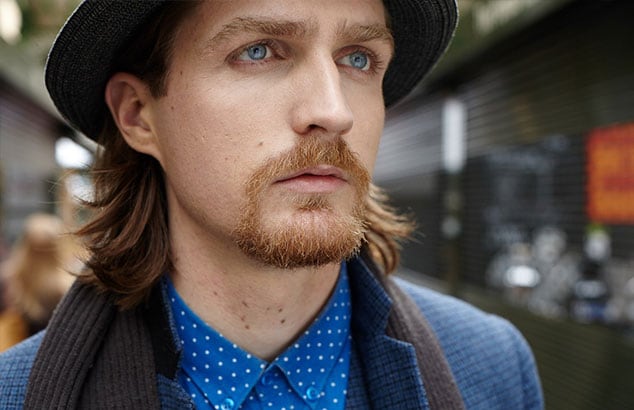 Man with shoulder long hair and henriquatre chin beard wearing a blue shirt, a suit jacket, and a hat.