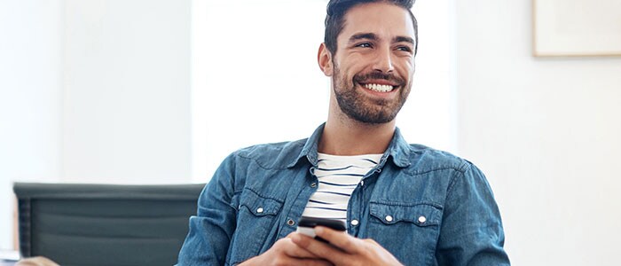 A man with beard stubble wearing a shirt holds a phone while smiling.